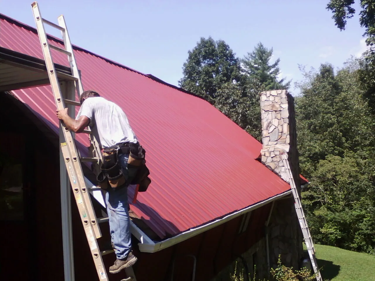 Roofer on a ladder beside a red metal roof and stone chimney — Soffit & Fascia Repair by Roofing Brothers in Boulder